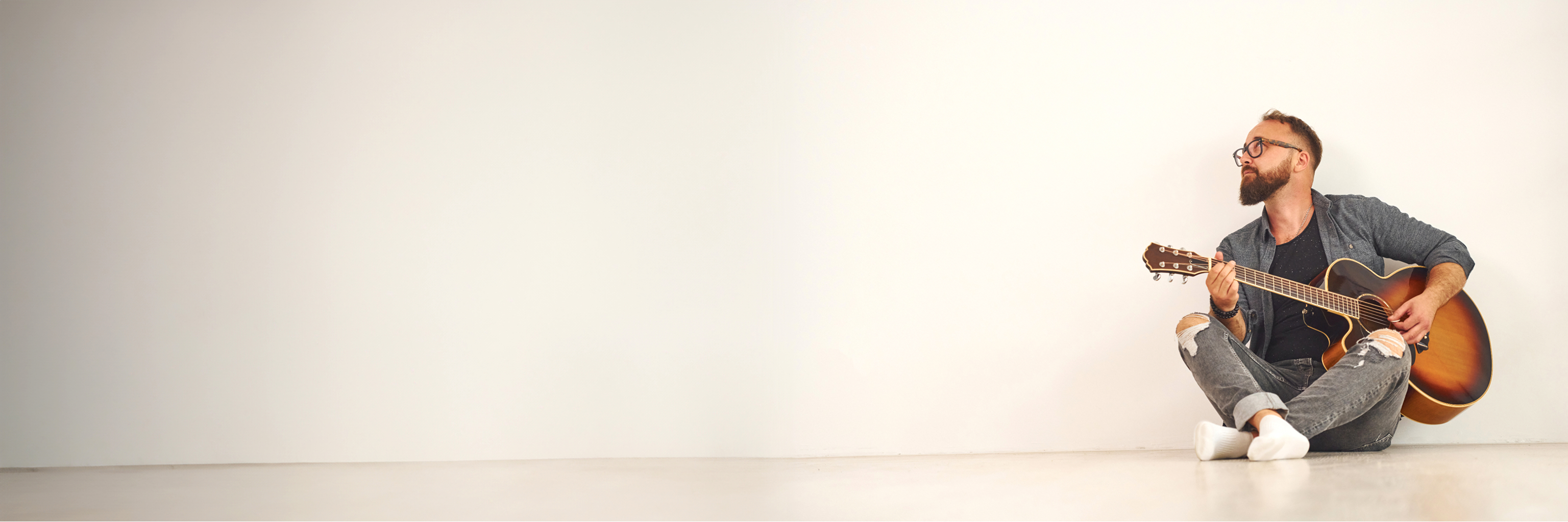 Wide image of a man sitting on the floor, playing the guitar in front of a white background
