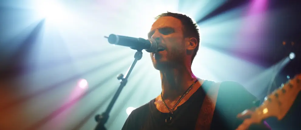 A man on stage performing live, singing into a microphone and playing guitar