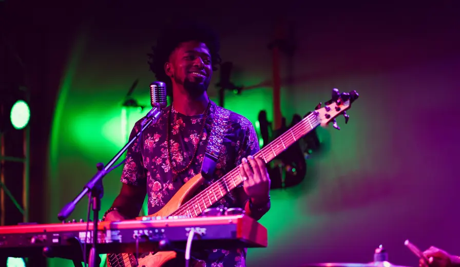 A man playing guitar on stage with a microphone and keyboard in front of him, moody colourful lighting