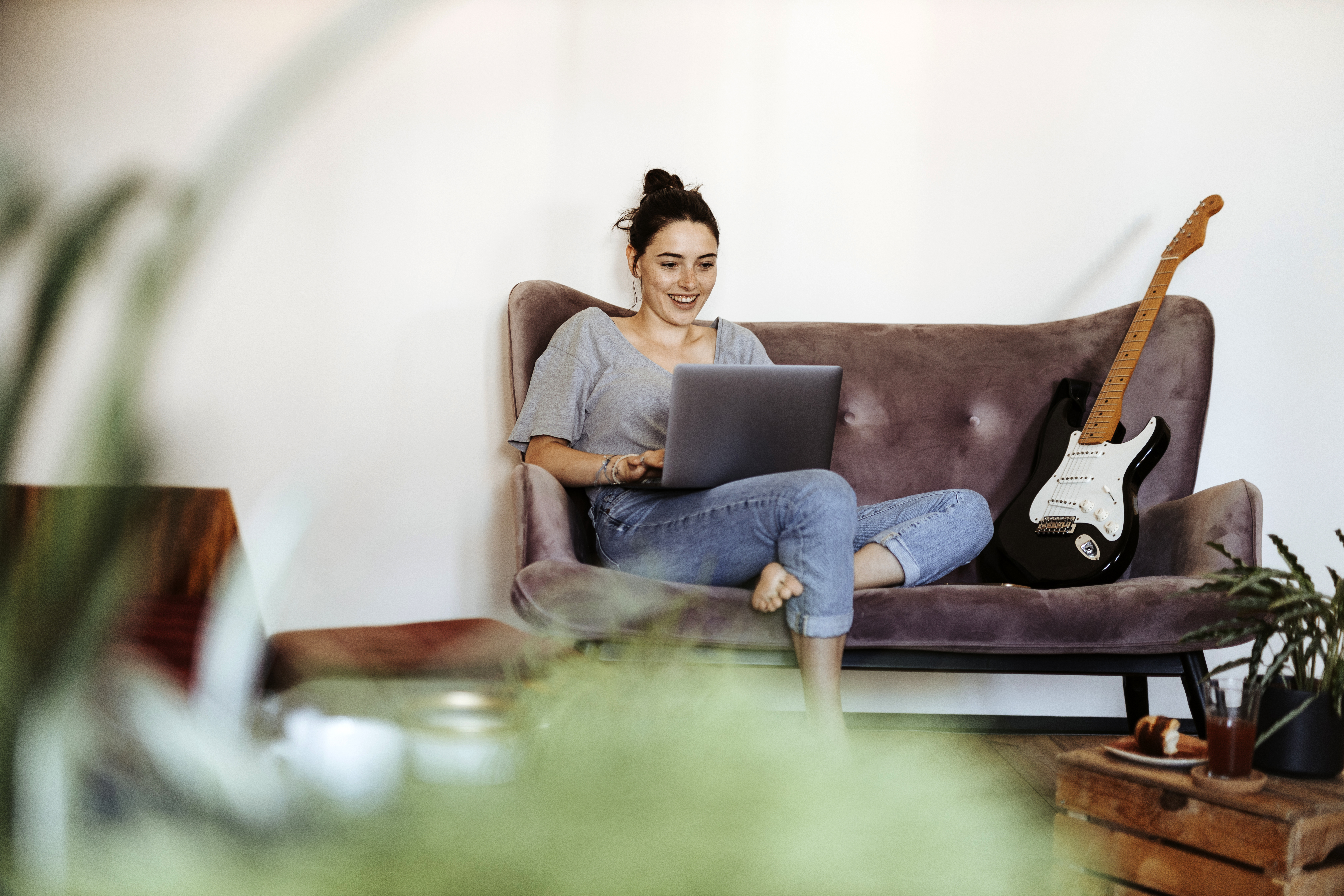 A young woman using a laptop sitting on a sofa with a guitar next to her