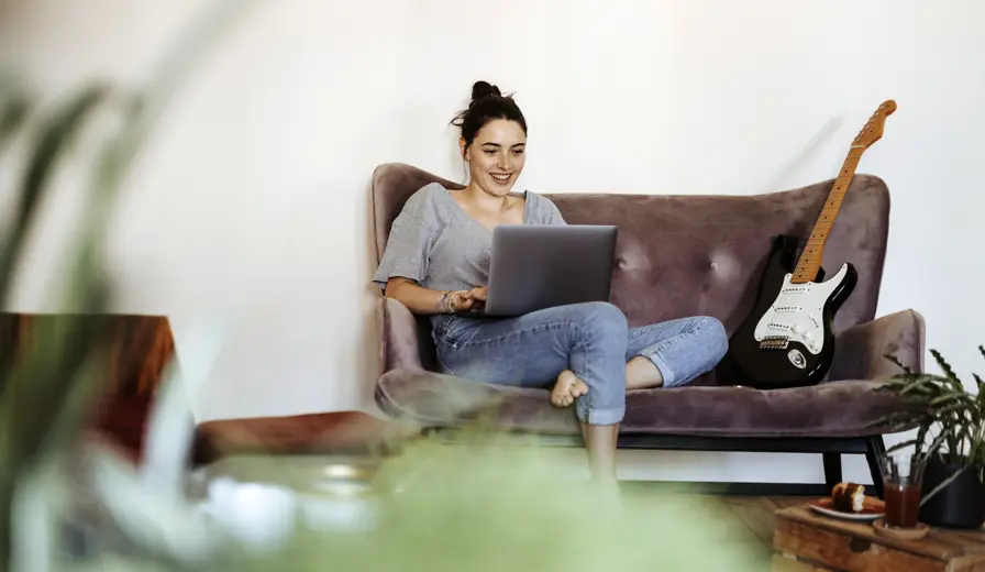 A young woman using a laptop sitting on a sofa with a guitar next to her